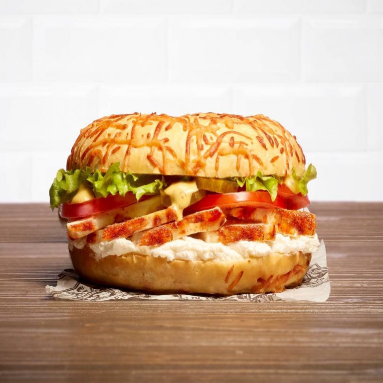 Un bagel avec du poulet, cream cheese, laitue, tomate, cornichons, sauce miel mayo, sur une table en bois et devant un mur en carrelage blanc, style métro.