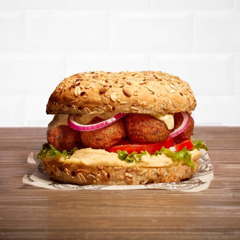 Un bagel végétalien aux falafels, tomate, oignon, houmous, sauce à la menthe sur une table en bois et devant un mur en carrelage blanc, style métro.