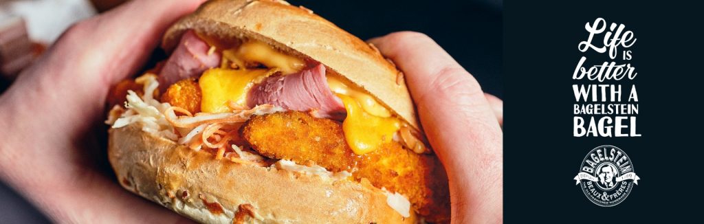 Gros plan d'une personne tenant un bagel farci de jambon, d'ingrédients croquants et de fromage fondu. L'arrière-plan présente le texte suivant : « La vie est meilleure avec un bagel Bagelstein. »